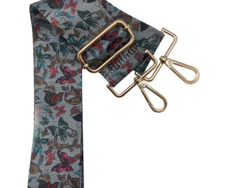 Pode incluir: Uma alça de tecido com padrão cinza e branco com fechos de metal dourado. A alça tem um design floral e de borboleta.
