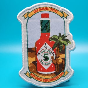 May include: Embroidered patch featuring a red hot sauce bottle with the text "Battling Bastards" and "Hot Sauce Deployment 2023." The patch includes the text "USS Bataan (LHD 5)" and a desert scene with a camel and palm tree.