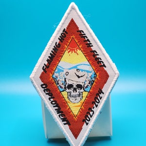 Könnte beinhalten: Ein rautenförmiger Aufnäher mit weißen, roten, gelben und blauen Farben. Der Aufnäher zeigt den Text "FLAMING HOT FIFTH FLEET DEPLOYMENT 2023-2024" um ein zentrales Design aus Schädel und Flammen.