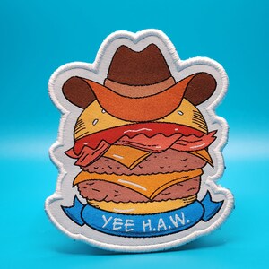 Może przedstawiać: Haftowana naszywka przedstawiająca kreskówkowego burgera w kapeluszu kowbojskim. Burger ma warstwy mięsa, sera i bekonu. Niebieski baner na dole głosi "YEE H.A.W." białymi literami. Naszywka ma białą obwódkę.