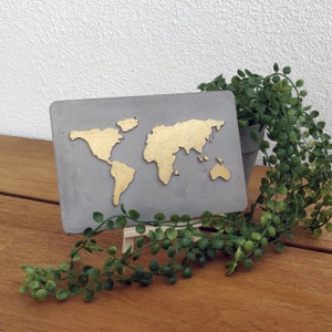 Könnte beinhalten: Eine graue Betonplakette mit einem goldenen Weltkarten-Design auf einer Holzoberfläche. Die Plakette steht auf einem kleinen Holzständer und eine grüne Pflanze befindet sich im Vordergrund.