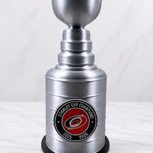 May include: A silver-coloured trophy cup featuring a tiered design and a circular emblem on the front. The emblem displays a red and black hurricane logo with the text "STANLEY CUP CHAMPIONS" and the years "2025" and "2026" printed in black.
