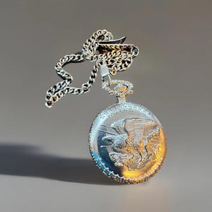 May include: Silver pocket watch with a chain. The watch face features a detailed relief of an eagle. The watch has a silver chain and a decorative crown at the top. The watch is suspended against a neutral background.