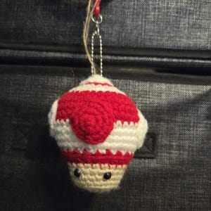 May include: A handmade crochet mushroom ornament in red, white, and beige. The mushroom has a red cap with white stripes, a beige stem, and black bead eyes. It hangs from a red clip and a silver chain.
