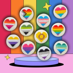 Puede incluir: Una colección de botones redondos blancos bordados a punto de cruz con varios diseños de corazón en colores arcoíris, rosa, azul, amarillo, verde y morado. Los botones se muestran sobre una plataforma azul sobre un fondo de arcoíris, con estrellas.