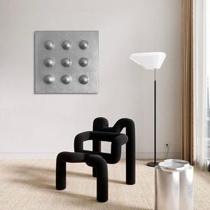 May include: An interior shot featuring modern furniture and decor. A black, sculptural chair with curved lines sits near a silver side table and a floor lamp. A square, silver wall art piece with nine raised circles hangs on the wall.