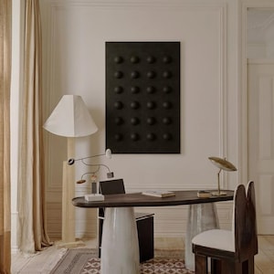 May include: An interior design scene featuring a dark wood desk with a white ceramic base and a metal base. A black wall art piece with raised circular elements hangs above the desk. A wooden chair and a floor lamp with a white shade are also visible.