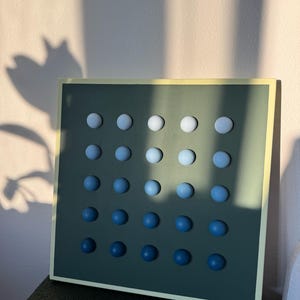 May include: A square wall art piece with a dark green background and a light yellow border. It features a grid of 25 evenly spaced, spherical, blue gradient spheres. The spheres transition from white at the top to dark blue at the bottom.