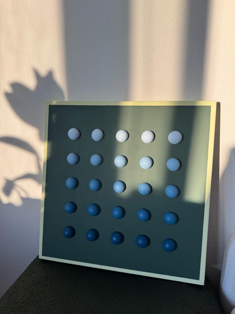 May include: A square wall art piece with a dark green background and a light yellow border. It features a grid of 25 evenly spaced, spherical, blue gradient spheres. The spheres transition from white at the top to dark blue at the bottom.