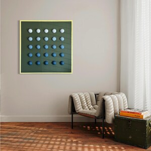 May include: A framed artwork featuring a grid of gradient blue spheres against a dark green background. The spheres transition from white to deep blue. The frame is a light gold color. The artwork hangs on a neutral wall above a chair and trunk.