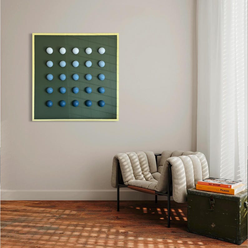 May include: A framed artwork featuring a grid of gradient blue spheres against a dark green background. The spheres transition from white to deep blue. The frame is a light gold color. The artwork hangs on a neutral wall above a chair and trunk.