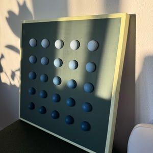 May include: A square wall art piece with a dark teal background and a grid of 25 wooden spheres. The spheres transition in color from white to dark blue. The frame is a light yellow color.