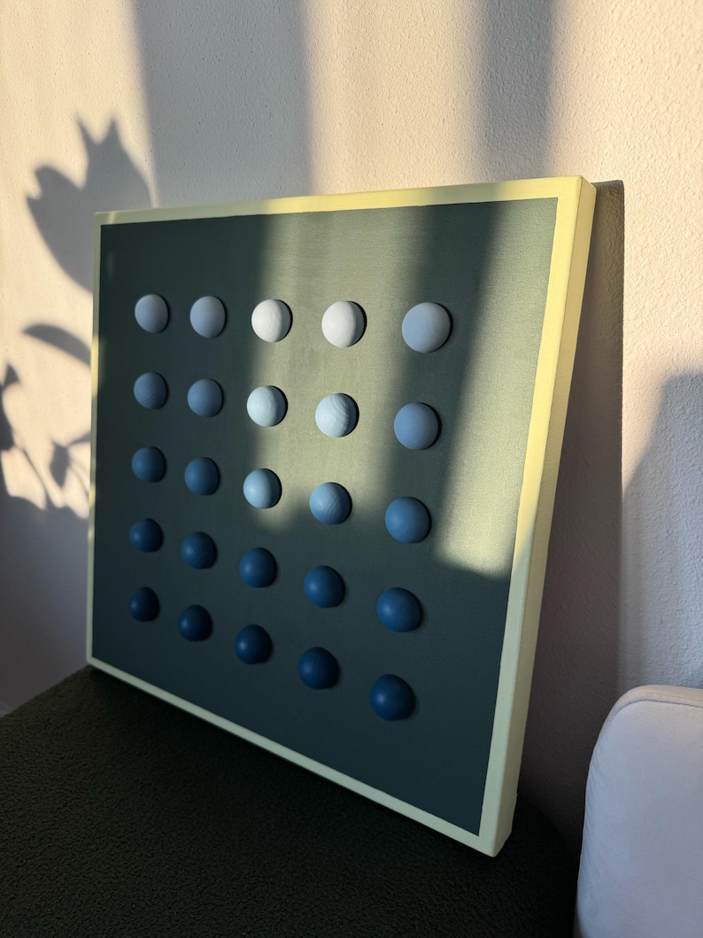 May include: A square wall art piece with a dark teal background and a grid of 25 wooden spheres. The spheres transition in color from white to dark blue. The frame is a light yellow color.