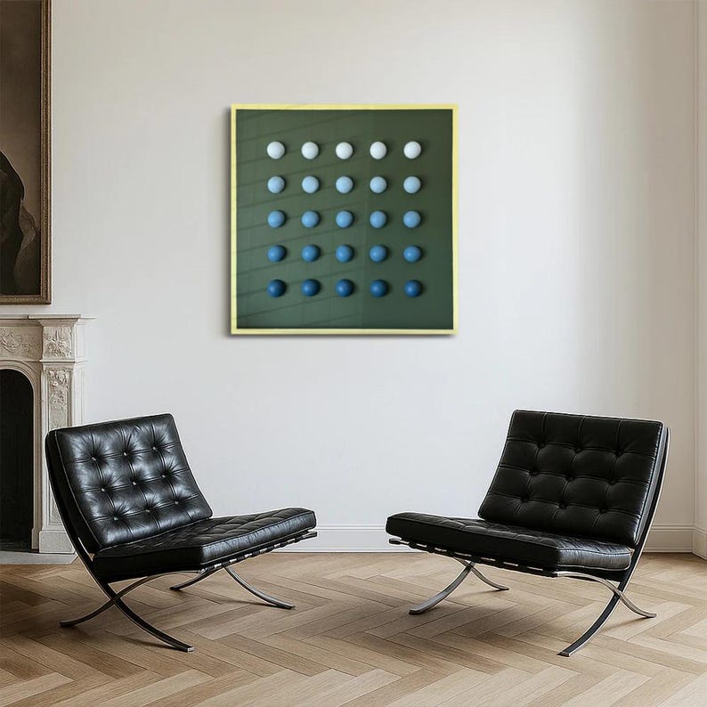 May include: An abstract art piece featuring a grid of gradient blue and white spheres on a dark green background. The artwork is framed in a light yellow border and hangs on a white wall. Two black leather chairs with chrome frames are in the foreground.