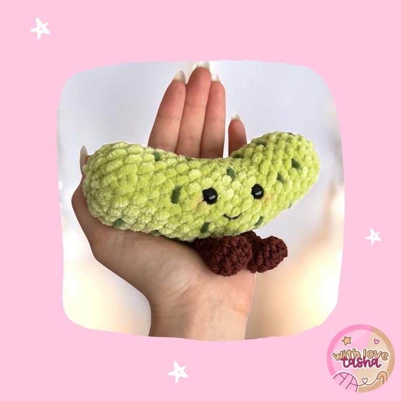 May include: A handmade, light green crocheted pickle with dark green spots, black eyes, and a smiling mouth. The pickle has brown feet and is held in a hand against a pink background with stars.