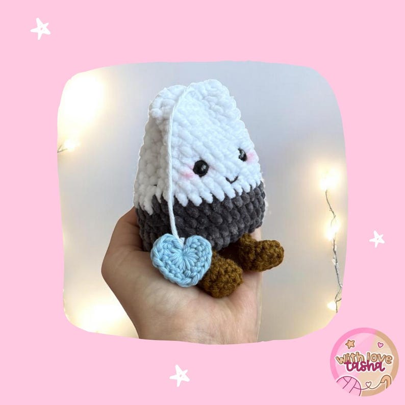 May include: A crocheted tea bag toy with a smiling face, a light blue heart tag, and brown feet. The tea bag is white on top, grey in the middle, and has a white string. A charming, handmade item.