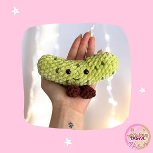 May include: A handmade, plush, green pickle with dark green spots, black eyes, and a smiling face. It has a small, brown bow tie. The pickle is held in a hand against a pink background with stars.