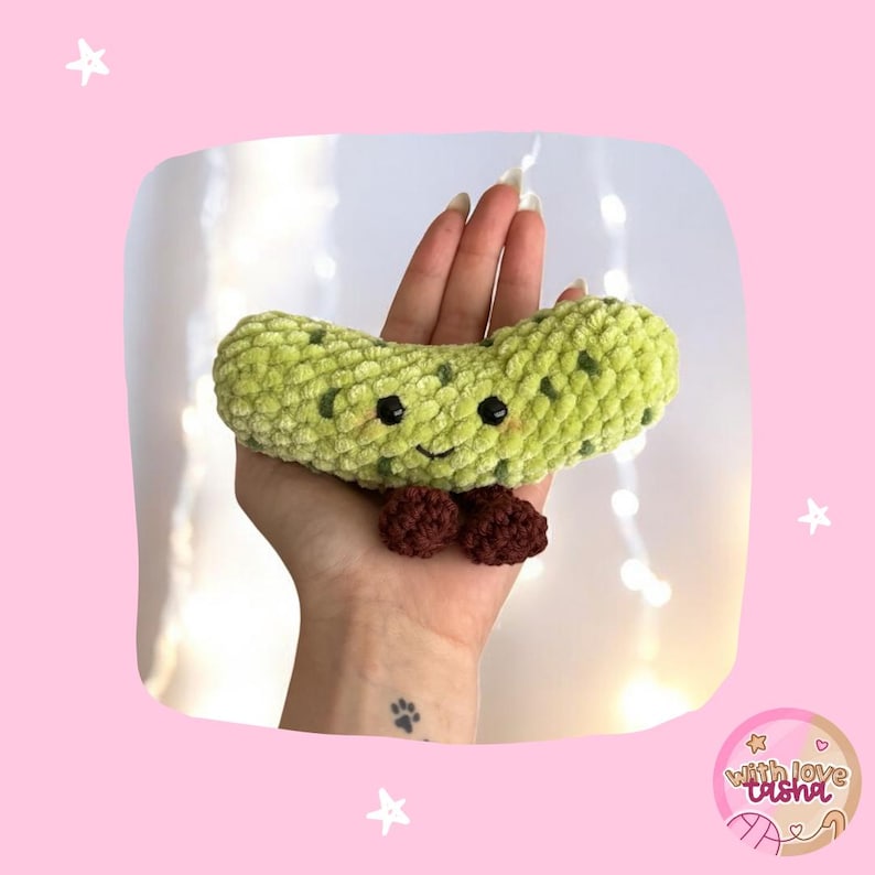 May include: A handmade, plush, green pickle with dark green spots, black eyes, and a smiling face. It has a small, brown bow tie. The pickle is held in a hand against a pink background with stars.