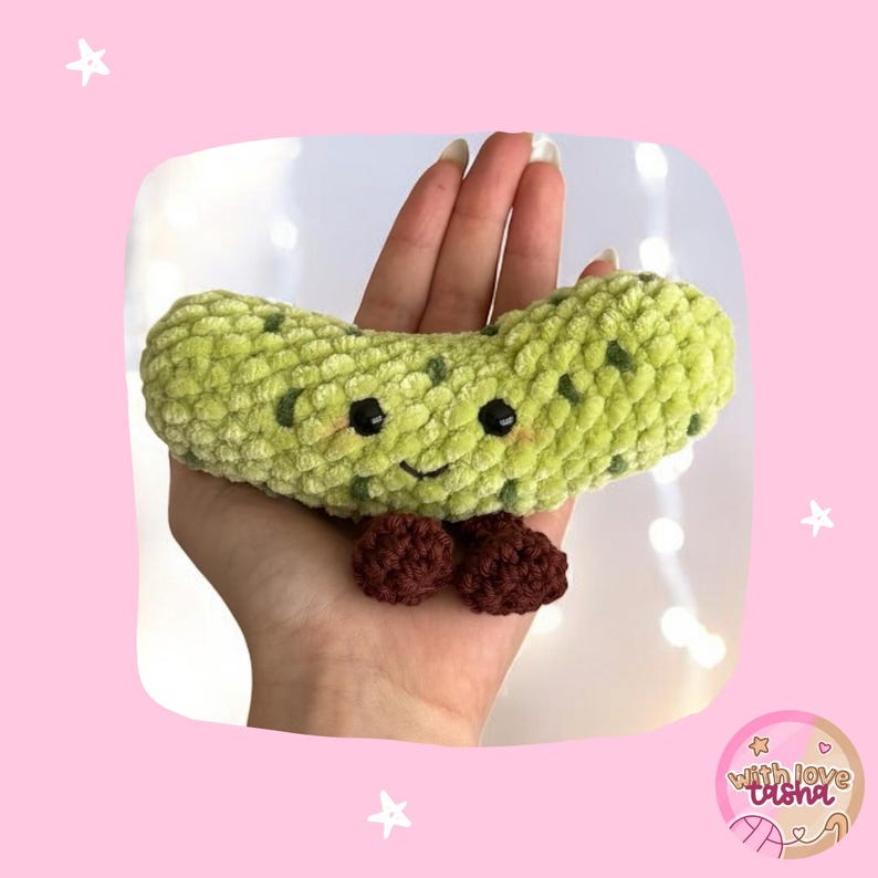 May include: A handmade, light green crocheted pickle with dark green spots, black eyes, and a smiling mouth. Two small, brown crocheted balls are attached at the bottom. The pickle is held in a hand against a pink background.