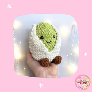 May include: A handmade crochet avocado plush toy with a smiling face, wrapped in a white shell. The avocado is light green, and the shell is white. The toy has brown feet.