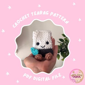 May include: A crochet teabag pattern PDF digital file featuring a white and dark gray plush teabag character with black eyes, a stitched smile, two brown feet, and a small turquoise heart tag attached by a white string, set against a pink background.