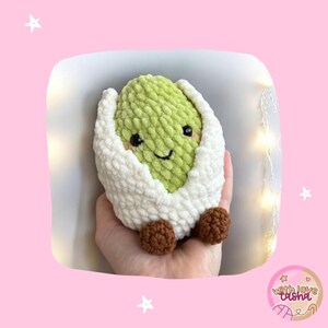 May include: A crocheted avocado toy with a smiling face, inside a white, fluffy eggshell. The avocado is green, and the eggshell is white. Brown feet are visible. The toy is held in a hand against a pink background.