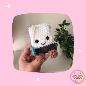 K&ouml;nnte beinhalten: Eine kleine, wei&szlig; geh&auml;kelte Marshmallow-Figur mit dunkelgrauer Basis, braunen F&uuml;&szlig;en und einem kleinen t&uuml;rkisfarbenen Akzent. Das Pl&uuml;schtier hat aufgestickte schwarze Augen und einen l&auml;chelnden Mund sowie eine weiche, texturierte Garnoberfl&auml;che.