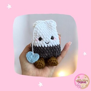 May include: A crocheted tea bag plush toy with a smiling face, light blue heart, and brown feet. The tea bag is white on top, grey in the middle, and has a string attached. The plushie is held in a hand.