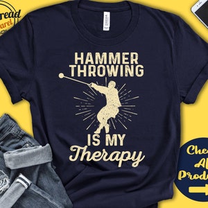 May include: A navy blue t-shirt with a white graphic of a person throwing a hammer. The text "Hammer Throwing is My Therapy" is printed in white on the shirt.