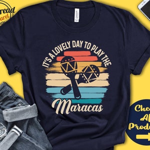 Pode incluir: Uma camiseta azul marinho com um gráfico de pôr do sol de estilo vintage e o texto "It's a lovely day to play the Maracas".