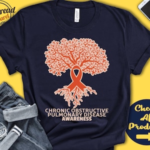 Puede incluir: Una camiseta azul marino con un diseño de árbol naranja. El árbol está hecho de muchos pequeños lazos naranjas. Las raíces del árbol están hechas de lazos naranjas. El texto "CHRONIC OBSTRUCTIVE PULMONARY DISEASE AWARENESS" está impreso debajo del árbol.