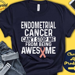 May include: A navy blue t-shirt with white text that reads "Endometrial Cancer Can't Stop Me From Being Awesome". A pink ribbon is below the text.