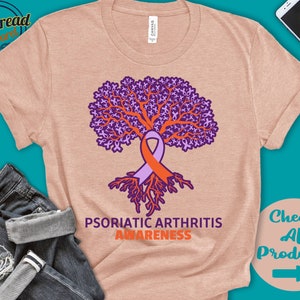 May include: A light pink t-shirt with a purple tree made of ribbons. The tree has orange roots and a purple and orange ribbon tied around the trunk. The text "Psoriatic Arthritis Awareness" is printed below the tree.