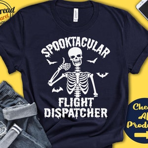 May include: Navy blue t-shirt with a white skeleton graphic giving a thumbs up, the words "Spooktacular Flight Dispatcher", and bats. The shirt also has the text "Check All Products" in a yellow circle.