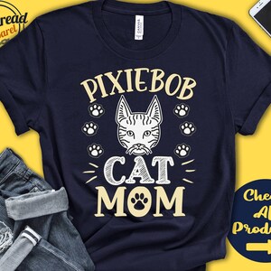 Puede incluir: Una camiseta azul marino con un diseño gráfico blanco que dice "Pixiebob Cat Mom" con una huella de pata y una ilustración estilizada de un gato.