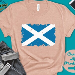 May include: A light pink t-shirt with a blue and white Scottish flag design. The flag is painted in a distressed style. The t-shirt is on a blue background with a pair of jeans and a cell phone.
