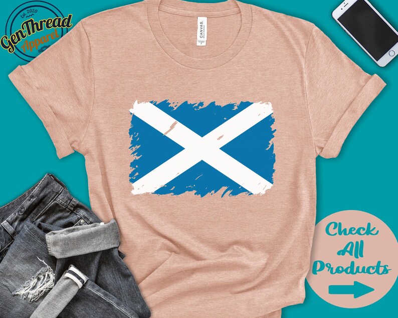 May include: A light pink t-shirt with a blue and white Scottish flag design. The flag is painted in a distressed style. The t-shirt is on a blue background with a pair of jeans and a cell phone.