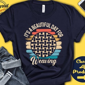 Peut inclure: Un t-shirt bleu marine avec un graphique rétro d'un panier tissé devant un coucher de soleil. Le texte "It's a beautiful day for weaving" est imprimé sur le t-shirt.
