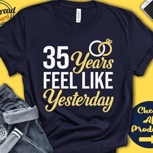 May include: Navy blue t-shirt with white text that reads "35 Years Feel Like Yesterday" and a gold ring with a diamond graphic. The shirt is on a yellow background with a pair of blue jeans and a phone.