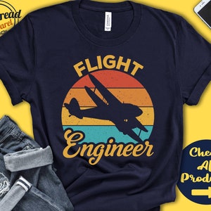 以下が含まれることがあります： 夕焼けを背景に飛行機が飛んでいるヴィンテージスタイルのグラフィックがプリントされたネイビーブルーのTシャツ。テキストは「Flight Engineer」。