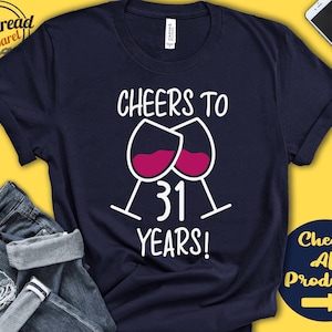 Puede incluir: Camiseta azul marino con el texto "CHEERS TO 31 YEARS!" y dos copas de vino llenas de vino tinto. La camiseta tiene cuello redondo y mangas cortas. Un par de vaqueros azules y un smartphone están en el fondo.