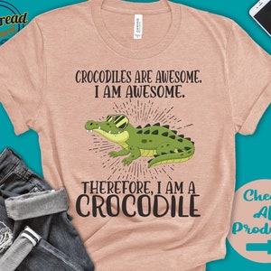 May include: A light pink t-shirt featuring a cartoon crocodile wearing sunglasses. The text on the shirt reads "Crocodiles are awesome. I am awesome. Therefore, I am a crocodile."