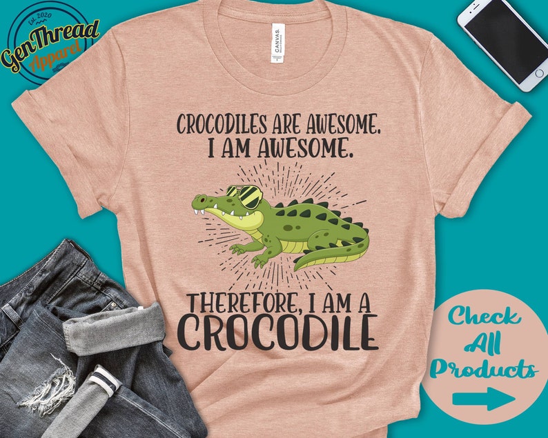May include: A light pink t-shirt featuring a cartoon crocodile wearing sunglasses. The text on the shirt reads "Crocodiles are awesome. I am awesome. Therefore, I am a crocodile."