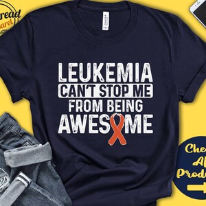 Può includere: Una maglietta blu navy con il testo "Leukemia Can't Stop Me From Being Awesome" in bianco con un nastro rosso.