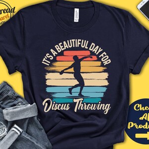 May include: A navy blue t-shirt with a vintage-style graphic of a discus thrower. The graphic features a sunset background with the text "It's a beautiful day for Discus Throwing".