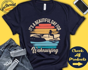 May include: A navy blue t-shirt with a vintage-style graphic that says "It's a beautiful day for wakesurfing". The graphic features a sunset with a woman wakesurfing behind a boat.