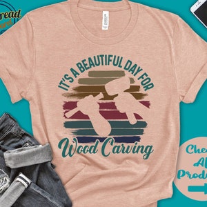 Puede incluir: Una camiseta rosa claro con un gr&aacute;fico de estilo vintage que dice "It's a beautiful day for wood carving". El gr&aacute;fico presenta una puesta de sol con una silueta de una persona tallando madera. El texto "Check All Products" est&aacute; en la esquina inferior derecha de la imagen.