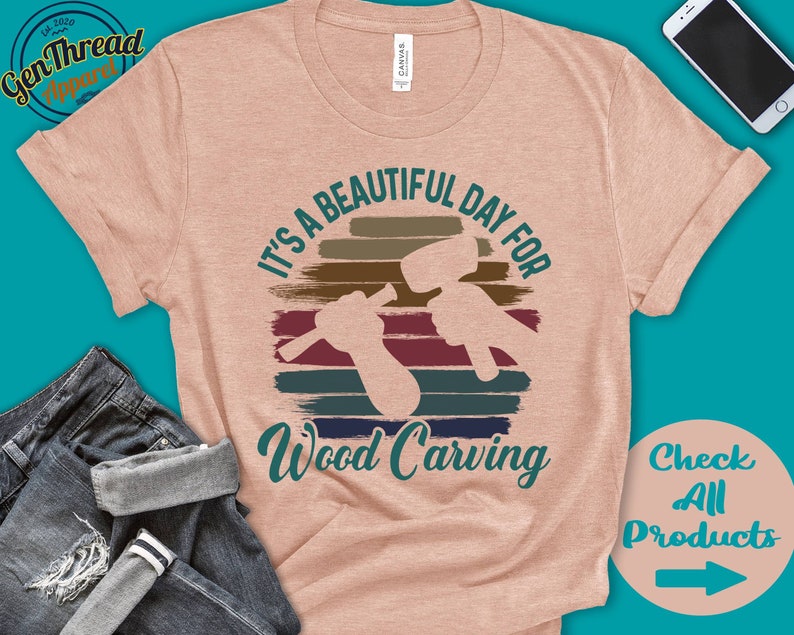 Puede incluir: Una camiseta rosa claro con un gr&aacute;fico de estilo vintage que dice "It's a beautiful day for wood carving". El gr&aacute;fico presenta una puesta de sol con una silueta de una persona tallando madera. El texto "Check All Products" est&aacute; en la esquina inferior derecha de la imagen.