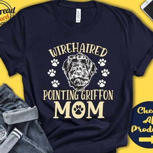 Peut inclure: Un t-shirt bleu marine avec le texte "Wirehaired Pointing Griffon Mom" en lettres blanches et jaunes. Une empreinte de patte est incluse dans le mot "Mom". Le design comprend également une illustration en noir et blanc d'un chien Wirehaired Pointing Griffon et des empreintes de pattes.