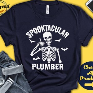 May include: Navy blue t-shirt with a white graphic of a skeleton giving a thumbs up, the text "Spooktacular Plumber", and bats. The shirt has a crew neck and short sleeves. The shirt is displayed with a pair of blue jeans.